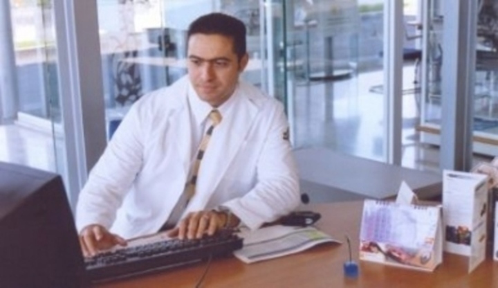 Muž sedící za stolem v bílém plášti s rukama na klávesnici počítače
